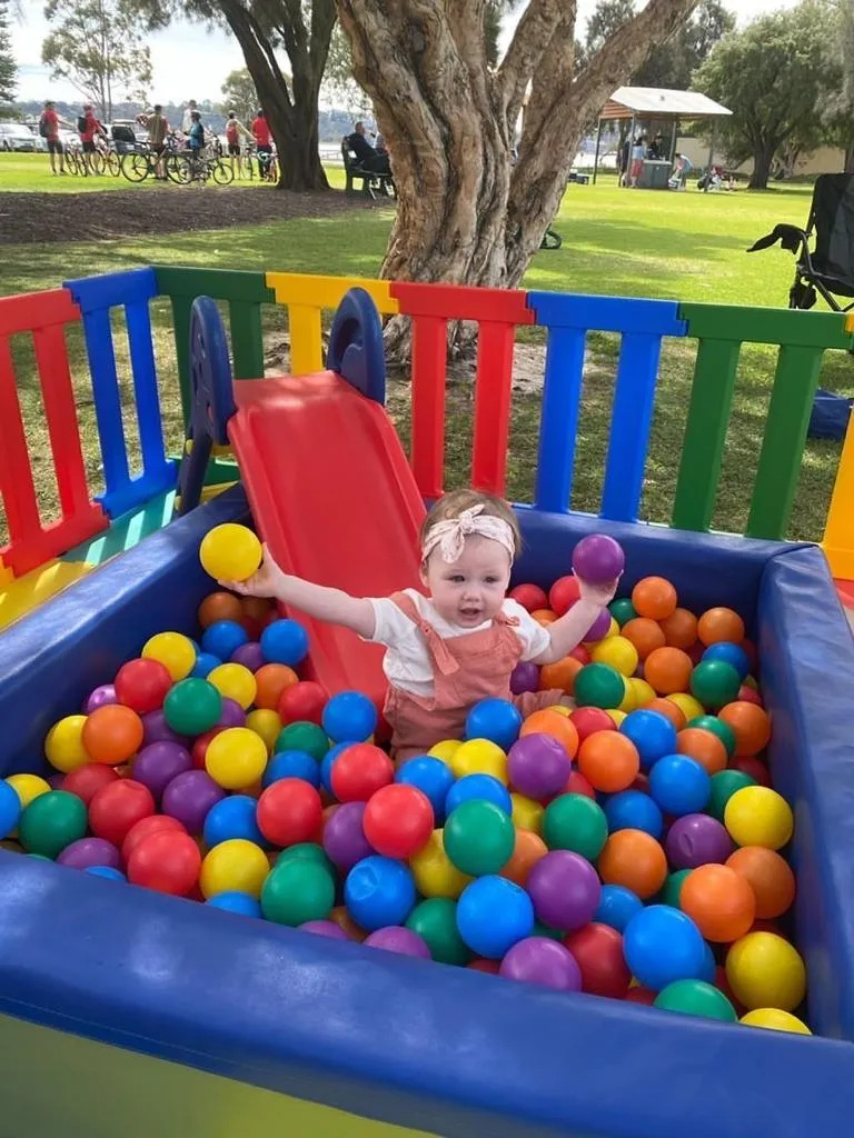 A toddler throwing coloured balls in a soft play ball pit with a red slide, set up in a park
